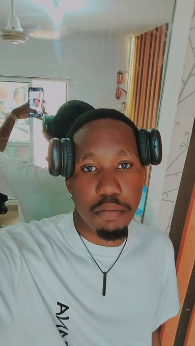 A man wearing headphones and a white shirt takes a selfie while someone behind him holds up a phone displaying his image.