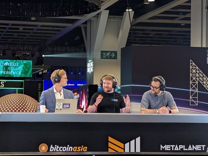 Three men wearing headsets sit at a broadcasting desk at a crypto event, with sponsor logos for 'bitcoin asia' and 'METAPLANET' visible on the front of the desk.