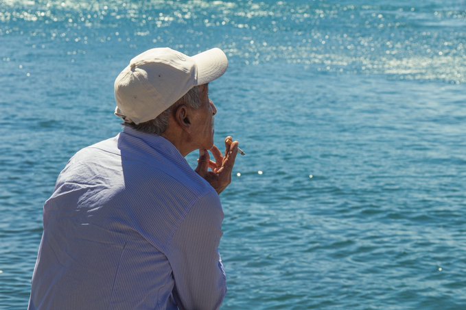 An older man in a beige cap and light blue shirt smokes a cigarette while gazing out over shimmering blue water.