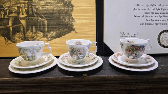 Three vintage floral-patterned teacups with matching saucers are displayed on a wooden surface against a backdrop featuring an Oxford college illustration and certificate.