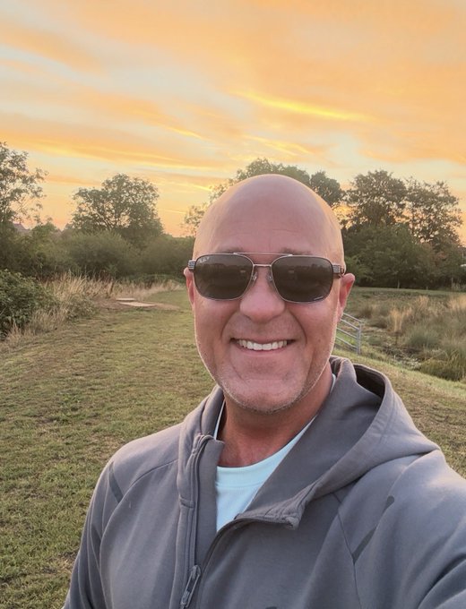 A bald man wearing sunglasses and a gray hoodie smiles for a selfie against a sunset sky with trees and grass in the background.