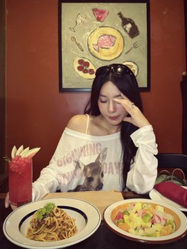 A woman with long dark hair wearing an off-shoulder 'COOKWARE' sweater sits at a restaurant table with pasta and salad dishes and a pink drink, beneath a framed picture of copper cookware.