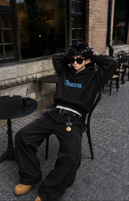 A person dressed in a black "Teana" hoodie, dark sunglasses, and baggy black pants sits relaxed in a chair at a sidewalk café, fitting the chill, positive vibes of a "gm frens" greeting.