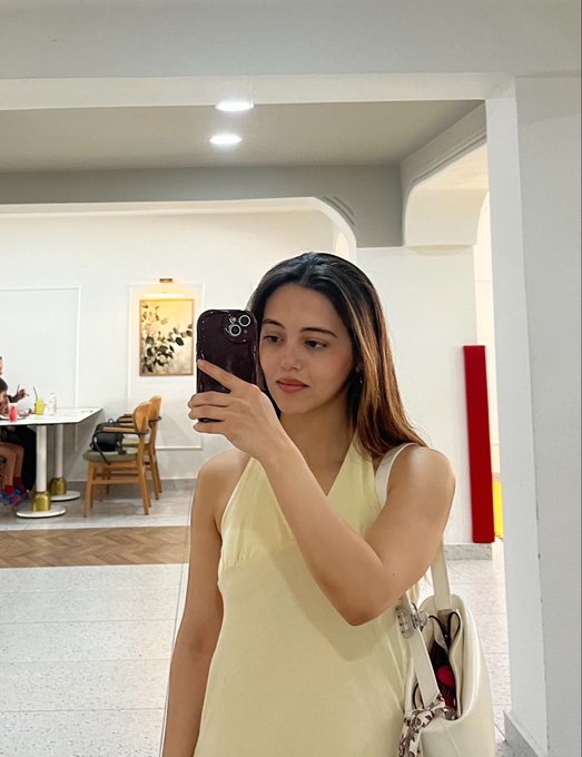 A young woman with long highlighted hair, wearing a pale yellow sleeveless dress, takes a mirror selfie with a smartphone in what appears to be a brightly lit cafe or salon.