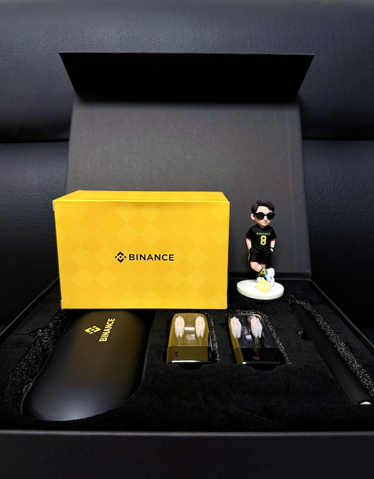 A Binance branded gift box set displayed in a black case, containing a black accessory case, two small boxes with what appear to be capsules, a pen, and a small figurine of a person wearing sunglasses and a number 8 shirt.