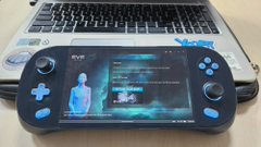 A handheld gaming device (looks like a PlayStation Portal) sitting in front of a laptop on a desk.