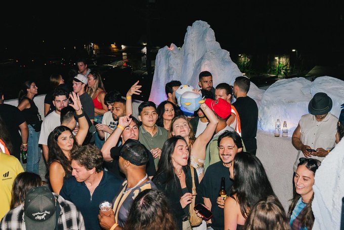 A crowded party scene at night with people socializing around ice-like sculptures, including someone holding a plush toy.