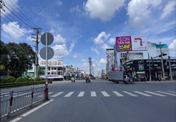 A daytime street view in Vietnam showing a wide intersection with motorbikes and a truck, flanked by commercial buildings with large billboards including an 'AEON TAN AN' sign and other Vietnamese advertisements.