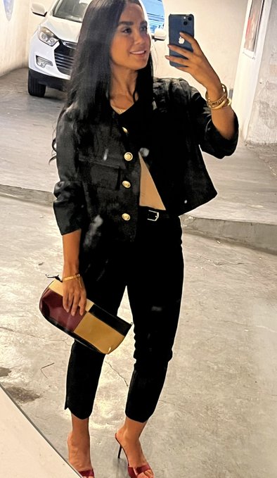 A mirror selfie of a woman with long dark hair wearing a black button-up jacket, black jeans, and red heels, holding a two-tone burgundy and tan handbag in what appears to be a parking garage.