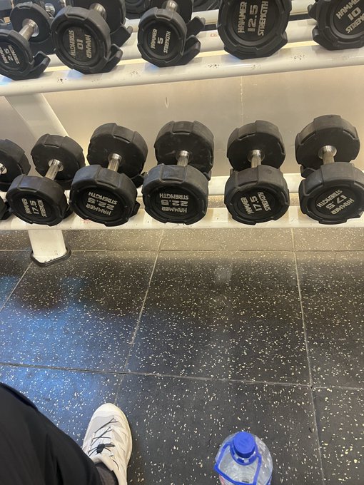 A pair of white sneakers stands in front of a rack of Hammer Strength dumbbells (ranging from 15 to 25 lbs) in a gym, with a water bottle nearby, capturing the moment of checking off a workout before the market opens.