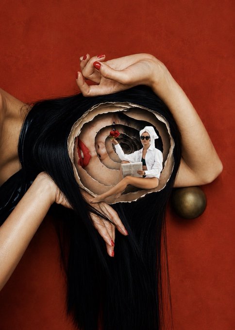 A surreal artistic photograph showing a woman with long dark hair lying down, where her hair forms a spiral portal containing a smaller figure of a woman reading and eating cherries.