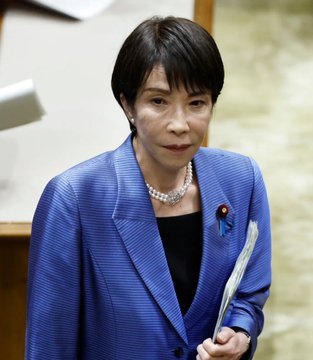 A woman in a blue pinstriped blazer with a pearl necklace and a red-and-blue ribbon pin holds documents, appearing serious. The image, paired with the post about Japan's economic decline, suggests a political figure addressing the country's financial crisis.