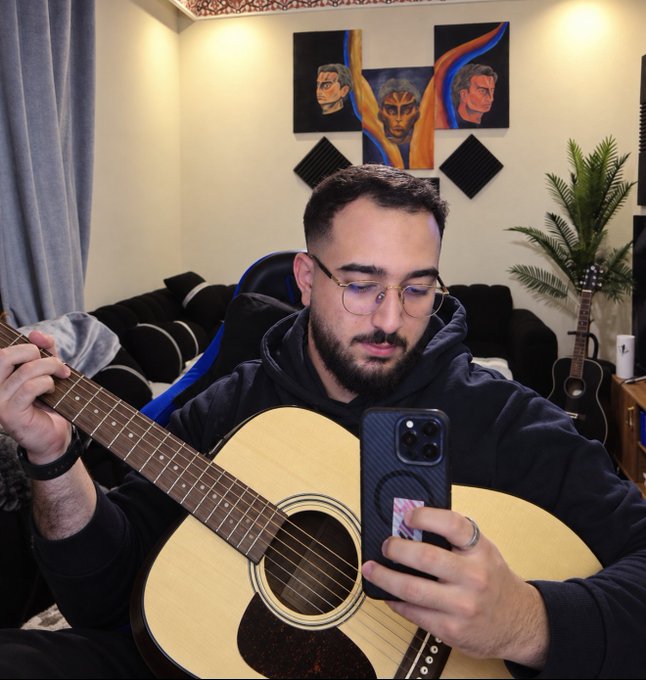A bearded young man wearing glasses and a black hoodie plays an acoustic guitar while looking at his smartphone, set against a backdrop featuring artistic paintings of faces and a second guitar leaning nearby. The image reflects the post's description of his creative passions, blending music and design in a personal, artistic setting.