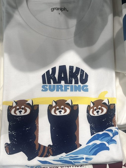 A folded white t-shirt by 'graniph' with a graphic design reading 'IKAKU SURFING' featuring three cartoon red pandas on a surfboard with blue waves.