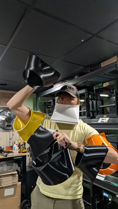 A person in a workshop wearing a LulzBot cap and a face shield, holding up multiple colorful 3D-printed objects, including black, yellow, and orange pieces.