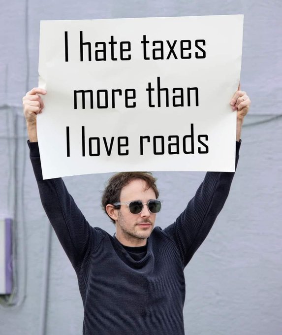 A man wearing sunglasses holds up a sign that reads 'I hate taxes more than I love roads' against a plain wall.
