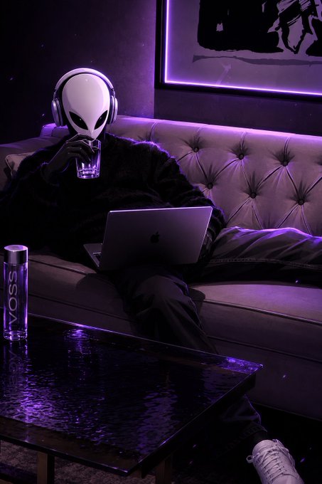 A person wearing a white alien mask, headphones, and a black fur coat sits on a tufted couch using an Apple laptop in a room lit with purple neon light, with a VOSS water bottle on the table.