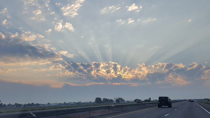 A scenic highway photograph capturing sunbeams streaming through scattered clouds at sunset, with a vehicle driving along the road toward the horizon.