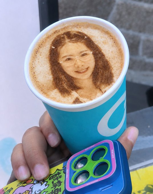 A hand holds a blue paper cup containing a coffee drink with latte art depicting a portrait of a smiling woman wearing glasses.