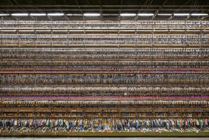 A photograph shows a vast warehouse filled with towering shelves densely packed with rolls of fabric in countless colors and patterns.