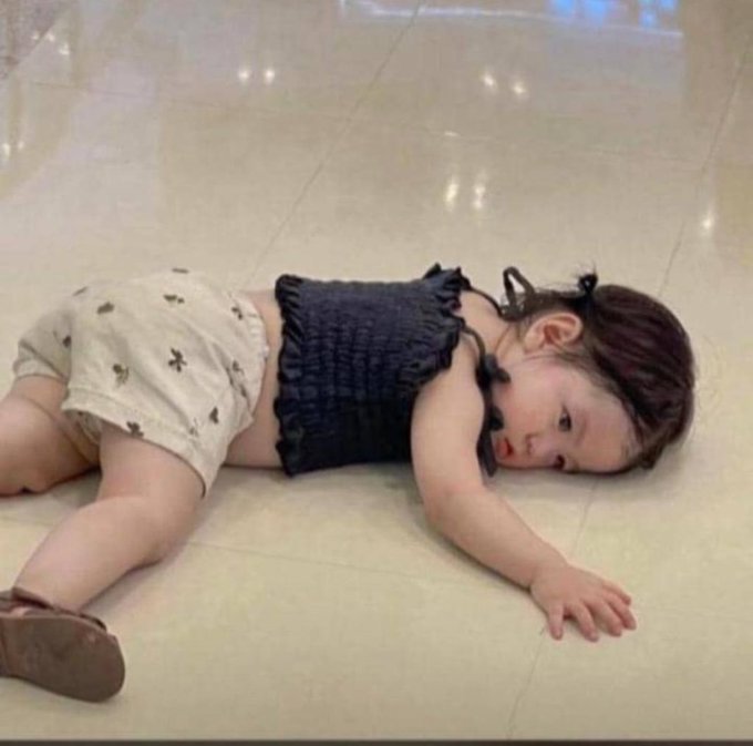 A young child wearing shorts and a dark top lies on a tiled floor, looking toward the camera.