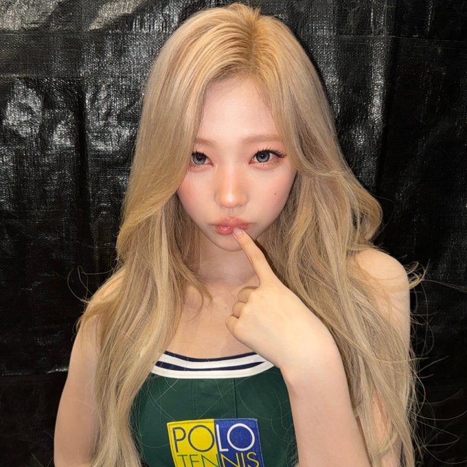 A selfie of a young woman with long blonde hair wearing a green and white 'POLO TENNIS' top, posing with her finger near her lips.