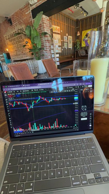 A laptop displaying a candlestick stock chart on a trading platform sits on a wooden table in a cafe setting.