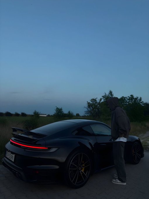 A person in a hoodie standing next to a black Porsche sports car at dusk in an open field.