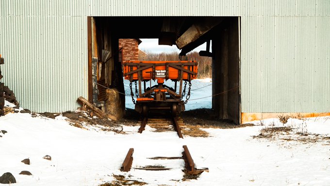 An abandoned industrial structure with an orange railway cart or hopper emerging from an opening, set against a snowy landscape with old tracks in the foreground.