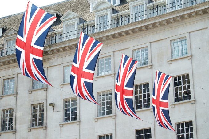 Four Union Jack flags hang on a line in front of a classical stone building, visually anchoring the UK focus of the Treasury's 54-firm tokenization task force, which includes BlackRock, Goldman Sachs, JPMorgan, and Morgan Stanley.