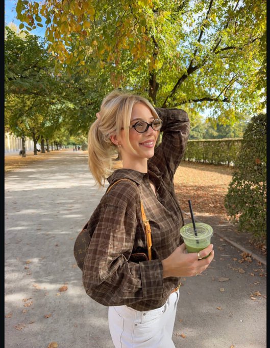 A young woman with blonde hair and glasses stands in an autumn park holding a green iced drink, wearing a brown plaid blouse and a Louis Vuitton bag.