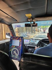 A view from inside a taxi in Hanoi, Vietnam, showing the driver and a digital advertisement display on the back of the front seat with passing street traffic visible through the windshield.