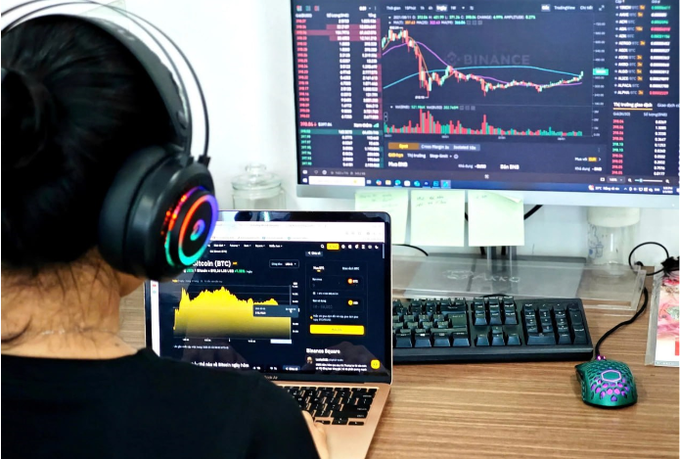 A person wearing RGB headphones sits at a desk using two screens, with a Binance trading chart visible on the monitor and a Bitcoin (BTC) chart on the laptop, alongside a pink gaming mouse.