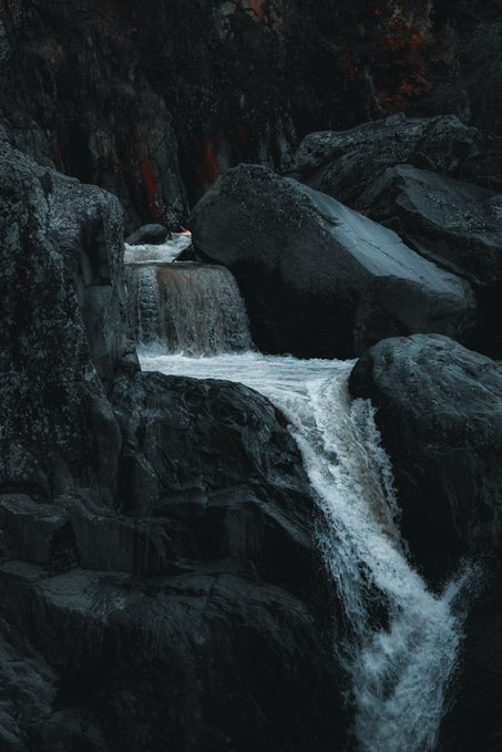 A dark, moody photograph of a small waterfall cascading over black volcanic boulders, with white water contrasting against the dark rock formations.