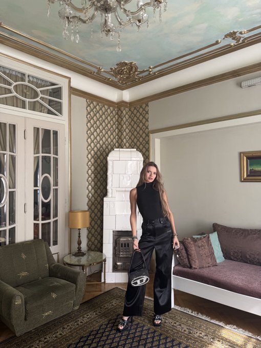 A woman in a black sleeveless top and black trousers stands in an ornate living room featuring a crystal chandelier, patterned ceiling, and vintage furniture.