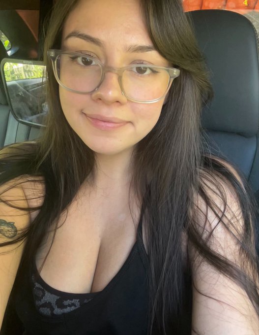 A close-up selfie of a young woman with long dark hair, clear-framed glasses, and a black tank top, sitting inside a vehicle.