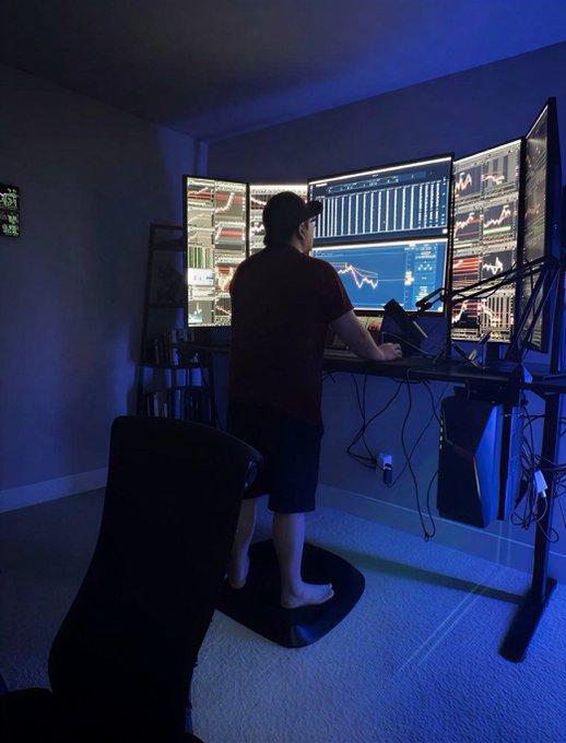 A person stands at a multi-monitor trading setup displaying various financial charts, candlesticks, and tickers under a blue-lit room, standing barefoot on an anti-fatigue mat while managing their $135 trading portfolio. The setup features several screens showing stock charts and technical analysis, reflecting a dedicated at-home trading workspace despite the modest portfolio size.