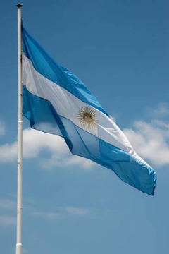 The light blue and white flag of Argentina with its central sun emblem waves against a blue sky with scattered clouds.