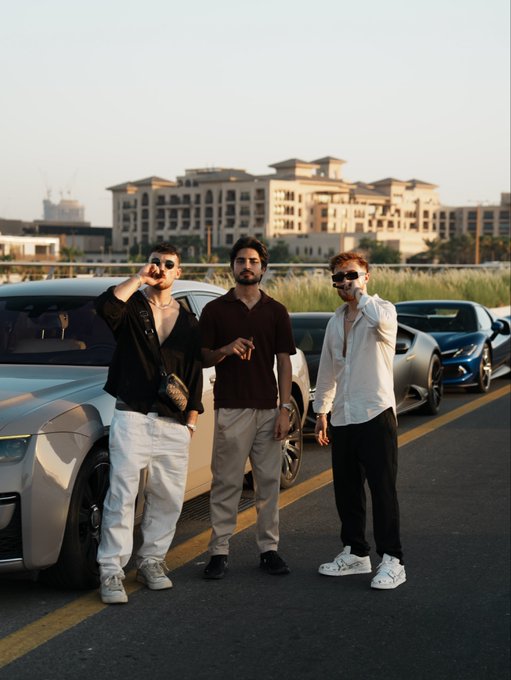 Three stylishly dressed men stand confidently in front of luxury sports cars, with an urban skyline in the background, responding to the "GM" (Good Morning) post with a vibrant morning vibe.