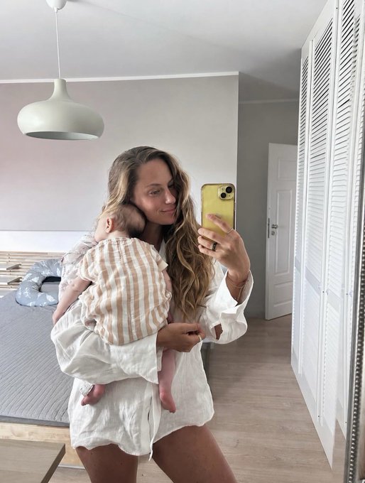 A woman takes a mirror selfie holding her sleeping baby in a striped onesie, both dressed in white tones in a bright bedroom. The caption "Bear market or not, I was heads down building" aligns with the image, portraying the dedication of building a business/family even during tough market times.