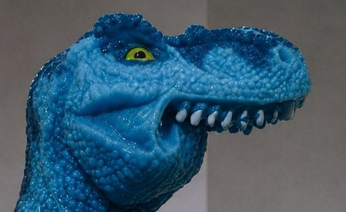 A close-up of a blue plastic T-Rex toy with a glossy, textured skin and bright yellow eye, showing sharp white teeth in a menacing grin. The dinosaur's predatory expression humorously mirrors the post's frustration about being locked out of the "crypto inner circle," as if stalking an elusive group while being unable to break in.