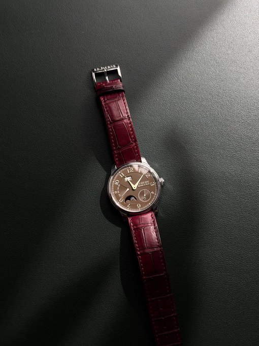 A wristwatch with a deep red leather strap and a brown dial featuring moon phase and other complications, lying on a dark, textured surface under directional light.