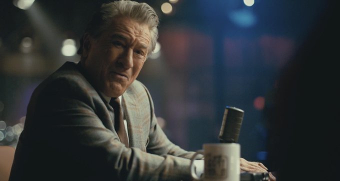 A cinematic shot of an older man with gray hair in a gray plaid suit, seated at what appears to be a bar or table with a microphone, suggesting a scene from a film.
