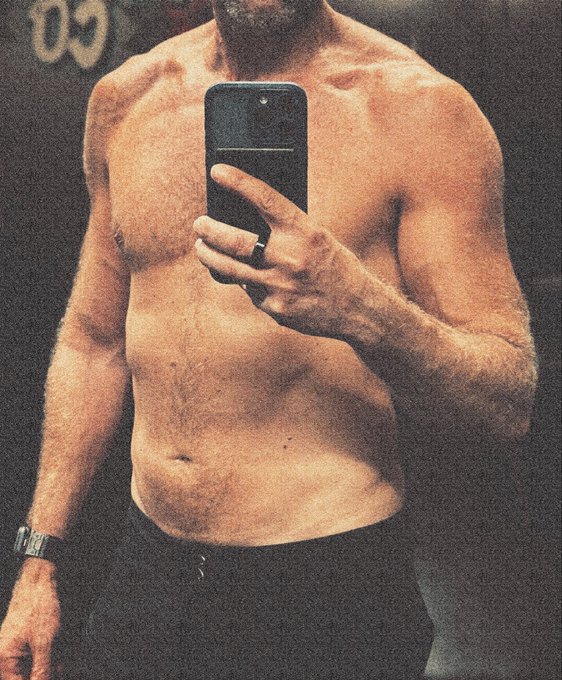 A shirtless, muscular man takes a mirror selfie in black shorts, holding a smartphone with the number "09" visible in the background, embodying the fitness and longevity goals of the post through his lean physique.