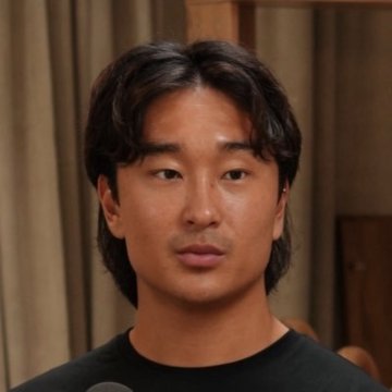 Portrait of a young Asian man with dark hair wearing a black shirt, photographed against a beige curtain background.