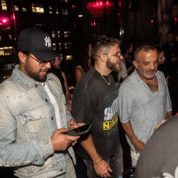 Three men, one wearing a New York Yankees cap, another in a black graphic t-shirt, and a third with a gray beard, navigate a crowded indoor venue with city lights visible in the background, reflecting the post's celebration of the "Doginal Dog community" gathering at DDNYC.