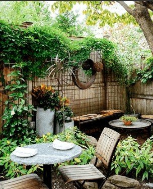 A lush garden patio features a small bistro table with white plates, surrounded by dense greenery, climbing vines, and a decorative circular mirror hanging on a bamboo fence.