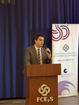 The image features a man standing behind a podium, giving a speech or presentation at an event. The man appears to be wearing a suit and tie, suggesting that he holds a professional role. There are two banners visible in the background, one of which is purple with white text reading "FECS". The other banner has a logo for a company named "Midnight Ntwrk".