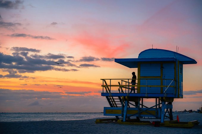 A blue and yellow lifeguard tower stands on a sandy beach under a pink and orange sunset sky.