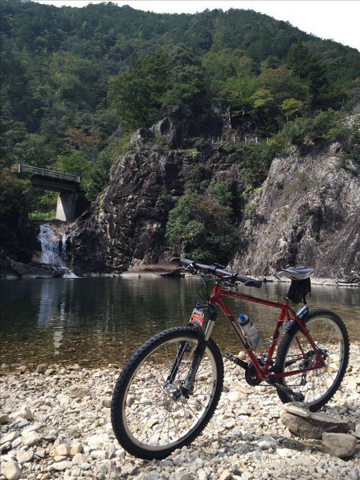 The image features a red mountain bike parked at the edge of a rocky shoreline, overlooking a river or lake. The scene suggests that someone has taken their bicycle for a ride along the water's edge, possibly enjoying the scenic views and natural beauty of the area.